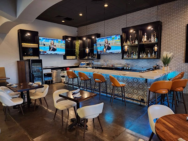 Stylish bar and restaurant interior with a patterned counter, orange barstools, dining tables, and large wall-mounted TVs.