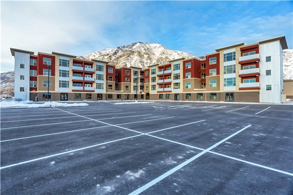 Newly constructed modern apartment complex with four floors, red and beige facade, and large parking lot with a mountain backdrop.