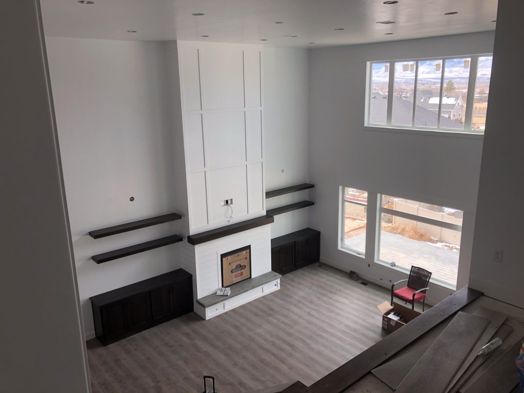 Spacious living room with a tall white fireplace, floating shelves, large windows, and wood-look flooring.