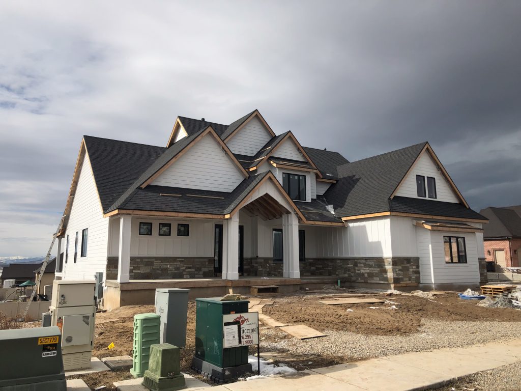 Two-story modern home under construction with white siding, dark roof, and stone accents, set against a cloudy sky.