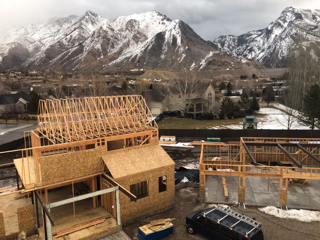 Residential home construction site with wooden framing, set against a backdrop of snow-covered mountains and a suburban neighborhood.