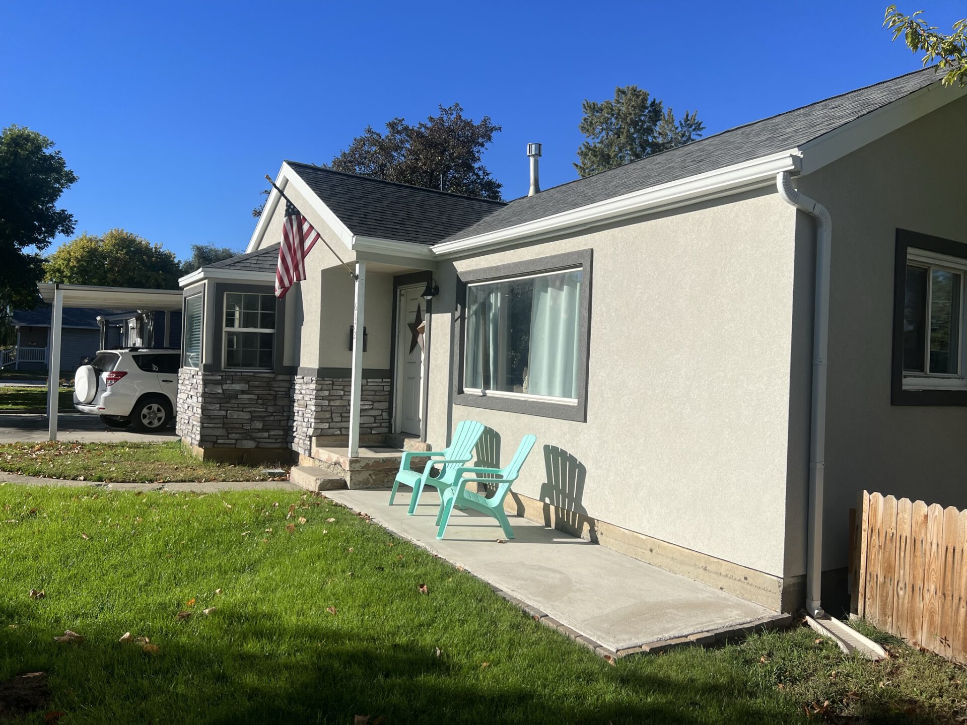 Single-story house with stone accents, American flag, green patio chairs, and a lawn, under a blue sky.